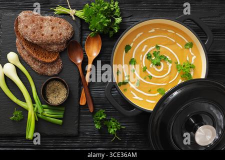 pannolini arrostiti e zuppa di carote in pentola nera su un tavolo di legno con cipolla verde, prezzemolo, pane di segale e cucchiai, vista orizzontale dall'alto, piano, no Foto Stock