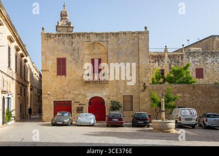 Mdina, Malta - 22 giugno 2021: Edificio storico in pietra con persiane rosse e auto parcheggiate in una piazza illuminata dal sole. Foto Stock