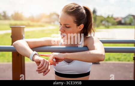 Adatta a una giovane donna che utilizza lo smartwatch per tenere traccia degli allenamenti nel parco della palestra all'aperto Foto Stock