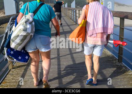 Due donne viste da dietro camminano su una passerella con sedie a sdraio e borse concetto di vacanza estiva Foto Stock