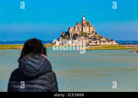 Una donna in primo piano, resa con un morbido bokeh, ammira Mont-Saint-Michel, la famosa isola di marea della Normandia sormontata dall'antica abbazia su un soleggiato... Foto Stock