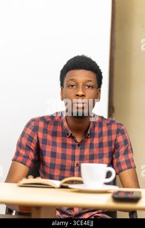 Giovane africano che studia a tavola con caffè e quaderno. Concetto: Stile di vita moderno degli studenti, produttività, apprendimento in caffetteria, preparazione accademica. Foto Stock