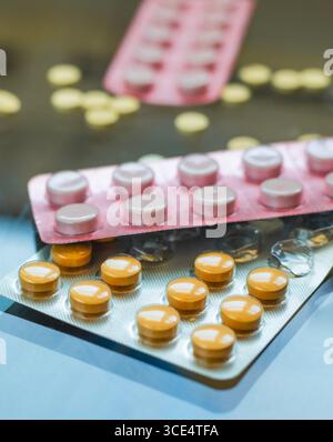 Le confezioni blister di varie pillole, incluse quelle arancioni e rosa, sono esposte contro una superficie riflettente. Alcune pillole gialle sfuse sono sparse in t Foto Stock