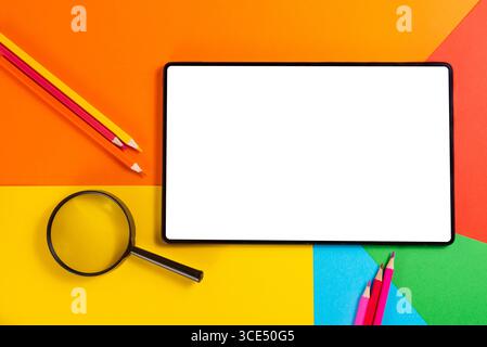 Tablet, lente d'ingrandimento e forniture scolastiche colorate su sfondo geometrico. Ideale per temi tecnologici e didattici. Foto Stock