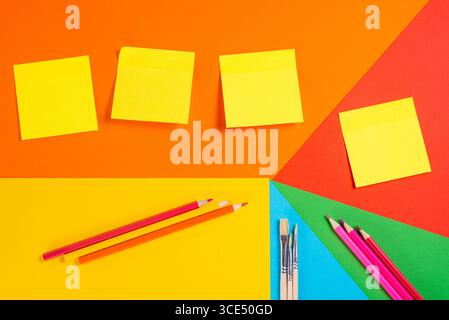 Note adesive, matite e penne disposte su sfondo geometrico colorato. Perfetto per la pianificazione e l'istruzione. Foto Stock