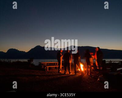 Amici in piedi intorno al falò dopo il tramonto con il lago Jackson in background, Colter Bay, il Parco Nazionale del Grand Teton, Teton County, Wyoming USA Foto Stock