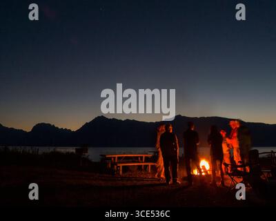 Amici in piedi intorno al falò dopo il tramonto con il lago Jackson in background, Colter Bay, il Parco Nazionale del Grand Teton, Teton County, Wyoming USA Foto Stock