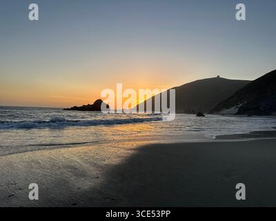 Tramonto dorato sull'Oceano Pacifico con le onde che scivolano sulla spiaggia sabbiosa e le scogliere aspre sullo sfondo. Foto Stock