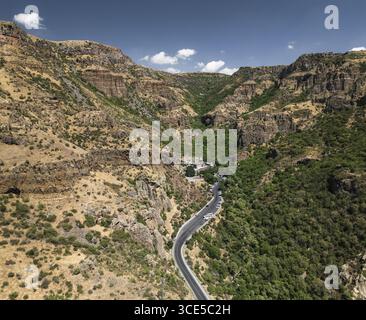 Veduta aerea di una strada tortuosa che attraversa una profonda gola, dove i pendii aridi e rocciosi incontrano un fitto e verde bosco, Goght, provincia di Ararat, Armenia. Foto Stock