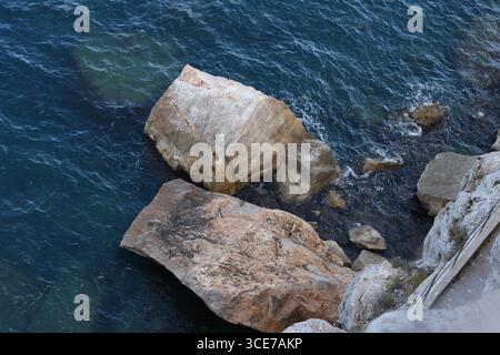 Enormi rocce si trovano ai margini del mare blu profondo, con onde che si infrangono delicatamente contro le loro superfici aspre. L'acqua rivela pietre sommerse. Foto Stock