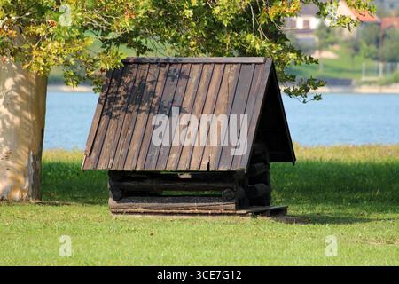 La struttura in legno A telaio Ad A si trova su una riva erbosa presso il lago blu, parzialmente ombreggiata da platano, offrendo posti a sedere rustici panoramici in un parco Foto Stock