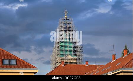 La torre della chiesa con cupola verde e intricata facciata è coperta da impalcature, che si innalzano su edifici con tetti di tegole rosse sotto spettacolari nuvole Foto Stock