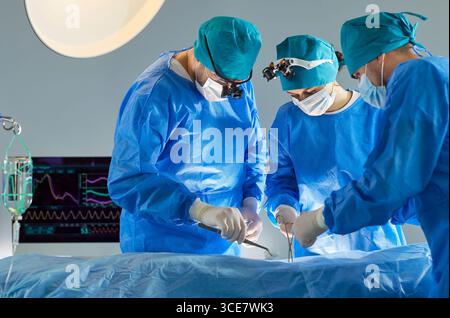 Team chirurgico in scrub e maschere che eseguono operazioni complesse sotto luci accese in camera sterile. Foto Stock