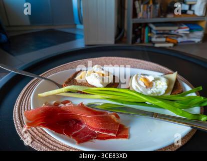 Due fette di pane tostato integrale ricoperte di uova in camicia, formaggio e cipolle verdi vengono servite insieme a fette di prosciutto stagionato su un piatto bianco, pronto Foto Stock