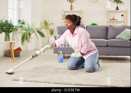 Giovane donna afroamericana che pulisce casa usando un moderno aspirapolvere senza fili Foto Stock