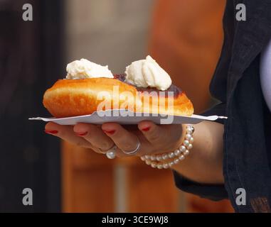 Le donne tengono a mano un vassoio di carta con due dolci fritti ricoperti di panna montata, braccialetto perle e unghie rosse visibili, fotografati in un ambiente all'aperto Foto Stock