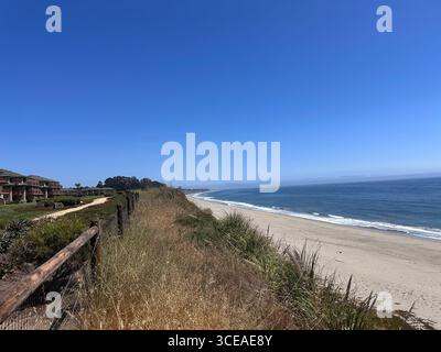Una vista panoramica della costa da un promontorio, che mostra dune erbose, case fronte oceano e un lungo tratto di spiaggia sabbiosa che incontra le calme acque blu Foto Stock
