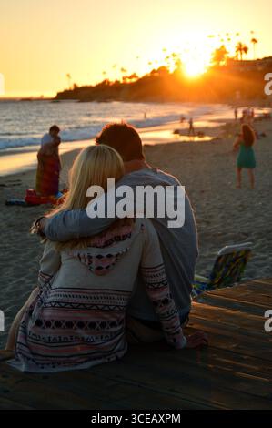 Una giovane coppia adulta condivide un romantico abbraccio mentre guarda il sole tramontare sull'Oceano Pacifico sulla riva di Laguna Beach in California Foto Stock