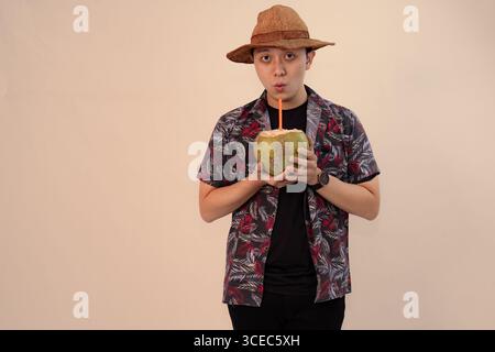 uomo in camicia stampata che regge noce di cocco con paglia arancione e indossa un cappello di paglia Foto Stock