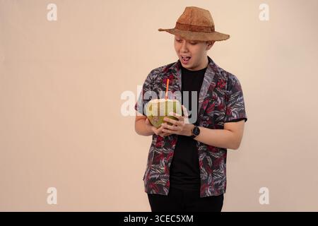 uomo in camicia stampata che regge noce di cocco con paglia arancione e indossa un cappello di paglia Foto Stock