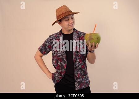uomo in camicia stampata che regge noce di cocco con paglia arancione e indossa un cappello di paglia Foto Stock