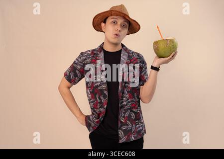 uomo in camicia stampata che regge noce di cocco con paglia arancione e indossa un cappello di paglia Foto Stock