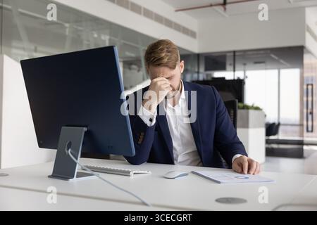 Giovane uomo d'affari stanco e infelice che tocca la testa sul posto di lavoro Foto Stock