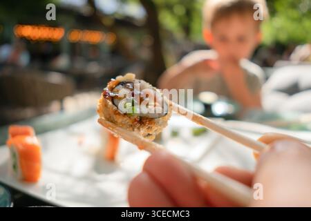 Primo piano di sushi roll al salmone al forno con bacchette, con un'autentica atmosfera da ristorante giapponese e un pasto in sottofondo per i bambini. Foto Stock