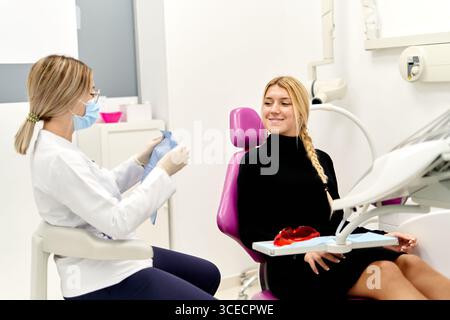 Una donna dentista che indossa una maschera si prepara ad una procedura in una moderna clinica odontoiatrica, mentre una giovane paziente si siede su una sedia dentale viola, awaitin Foto Stock