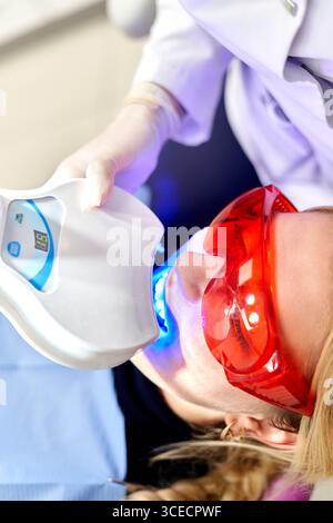 Un professionista di clinica dentale esegue una procedura di sbiancamento dei denti utilizzando attrezzature avanzate. Il paziente indossa occhiali protettivi durante il trattamento Foto Stock
