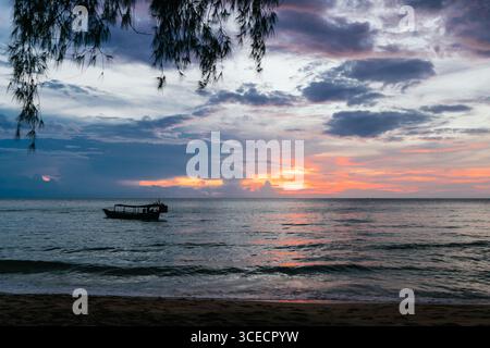 Un tramonto sereno sull'isola di Koh Rong, in Cambogia, con vivaci sfumature arancione e viola che si riflettono nelle acque calme. Sagome di alberi e di una barca Foto Stock