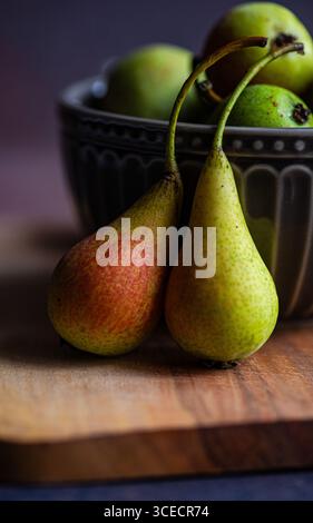 Primo piano di due pere selvatiche appoggiate su un asse di legno con una ciotola decorativa in ceramica riempita con altre pere sullo sfondo, che mostrano la loro naturale natura Foto Stock