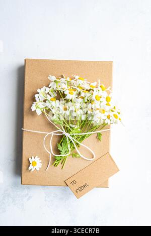 Vista dall'alto di un mazzo di margherite bianche fresche legate con cordoncino su una confezione regalo marrone con un cartellino bianco. Lo sfondo è una superficie bianca testurizzata, creato Foto Stock