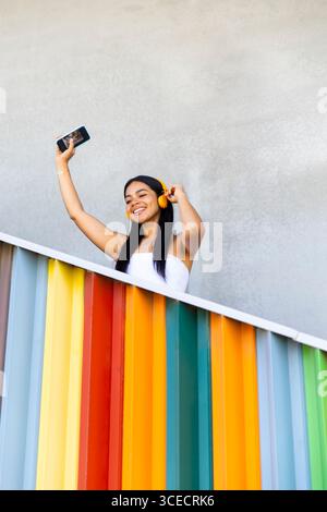 Una donna che indossa cuffie vivaci ama la musica mentre si trova sullo sfondo di un'architettura moderna e colorata. Ha uno smartphone, abbraccia un Foto Stock