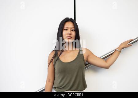 Una giovane donna cinese si appoggia contro una ringhiera, godendosi la calda giornata estiva in città. Indossa una canotta casual e guarda con sicurezza la t Foto Stock