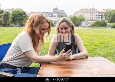 Una madre e una figlia felici si godono del tempo di qualità insieme in un parco, seduti a un tavolo di legno e sorridenti mentre condividono qualcosa su uno smartphone. Il Foto Stock