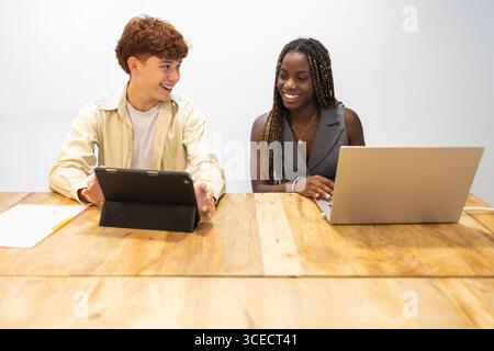 Un giovane team multirazziale collabora con entusiasmo in un ufficio di coworking. Un uomo usa un tablet mentre il suo collega lavora su un notebook, riflettendo Mo Foto Stock