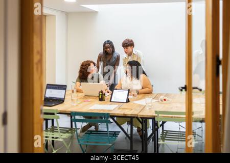 Un gruppo eterogeneo di colleghi impegnati in uno spazio di coworking, discutendo idee con notebook e tablet, promuovendo il lavoro di squadra e l'inclusività in una modalità Foto Stock