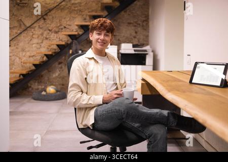 Un giovane uomo siede casualmente in un moderno spazio di coworking, tenendo in mano una tazza di caffè. Un tablet visualizza grafici nelle vicinanze, creando un ambiente di lavoro rilassato. Foto Stock