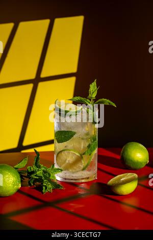 Un rinfrescante mojito classico decorato con lime e menta, abbinato a un audace piano da tavolo rosso con un caldo sfondo giallo. Perfetto per un cocktail estivo Foto Stock
