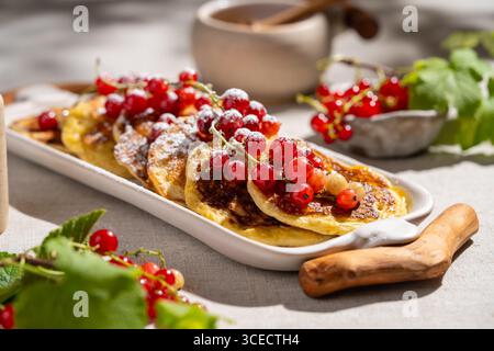 Deliziosi pancake adornati con ribes rosso sono serviti su un piatto di ceramica bianca. Poggiano su un tessuto beige testurizzato, creando una deliziosa colazione Foto Stock