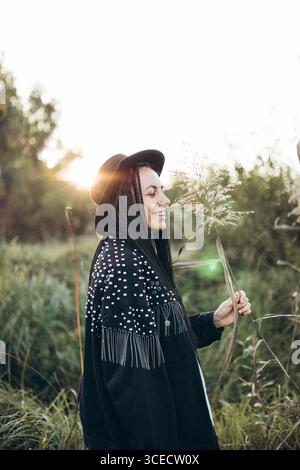Una donna con i capelli intrecciati e un ampio cappello a setole gode di un momento sereno nella natura durante il tramonto. Si erge in mezzo ad erba alta, bagnata dall'oro caldo Foto Stock
