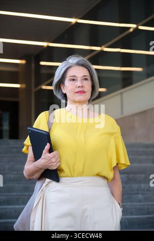 Donna matura in piedi con sicurezza all'aperto, indossa un elegante top giallo e regge un tablet. Trasuda professionalità e modernità in un ambiente urbano Foto Stock