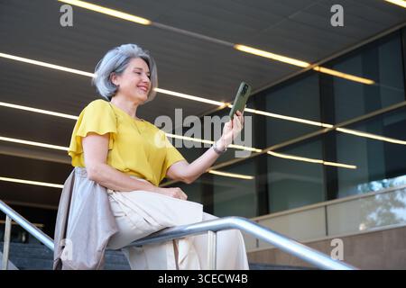 Una donna allegra e matura con una camicetta gialla scatta un selfie con il suo smartphone mentre è seduta su una ringhiera all'aperto. Sta sorridendo e si sta godendo la mamma Foto Stock