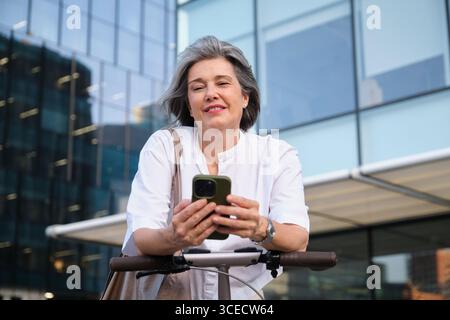 Una donna matura con un'elegante camicetta bianca sorride mentre usa uno smartphone all'aperto, appoggiandosi su uno scooter elettrico, con un moderno edificio in vetro nella b Foto Stock
