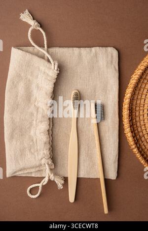 Vista dall'alto di due spazzolini da denti in bambù su un sacchetto di tessuto beige con cestello in vimini su sfondo marrone, per una vita ecocompatibile e senza sprechi Foto Stock