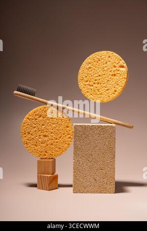 Lo spazzolino in bambù ecologico si appoggia su due spugne arancioni circolari. Un blocco di sughero rettangolare completa la disposizione su uno sfondo marrone minimalista Foto Stock
