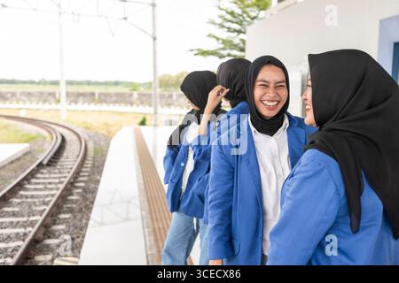 Un gruppo di donne musulmane asiatiche che indossano uniformi blu e hijab neri, condividono una risata mentre aspettano in una stazione ferroviaria, simboleggiando un allegro ritorno Foto Stock