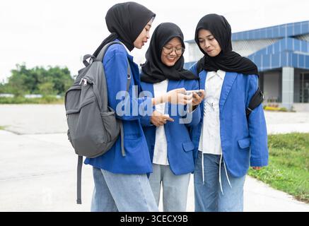 Tre studenti musulmani asiatici in uniforme blu si riuniscono felicemente fuori, condividendo un momento su uno smartphone. Sembrano pronti e desiderosi di tornare Foto Stock