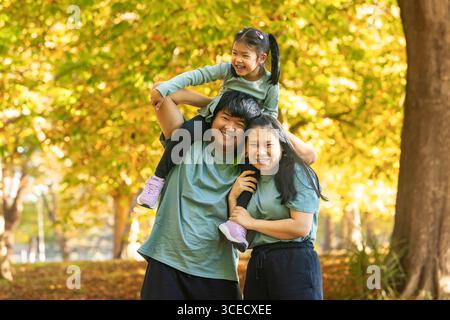 Una gioiosa famiglia asiatica gode di una giornata di sole nel parco, catturando preziosi momenti di legame. I genitori sorridono calorosamente mentre la figlia si siede sul loro Foto Stock
